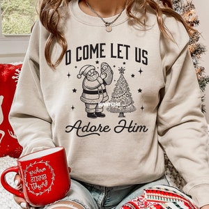 Pode incluir: Camisola bege com gola redonda com um gr&aacute;fico preto do Pai Natal segurando um beb&eacute; ao lado de uma &aacute;rvore de Natal. O texto diz "O Come Let Us Adore Him". Uma caneca vermelha com texto branco est&aacute; em primeiro plano.