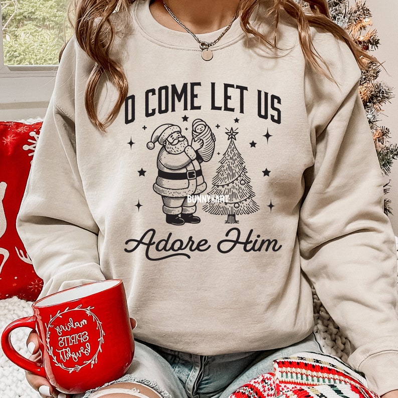 Pode incluir: Camisola bege com gola redonda com um gr&aacute;fico preto do Pai Natal segurando um beb&eacute; ao lado de uma &aacute;rvore de Natal. O texto diz "O Come Let Us Adore Him". Uma caneca vermelha com texto branco est&aacute; em primeiro plano.