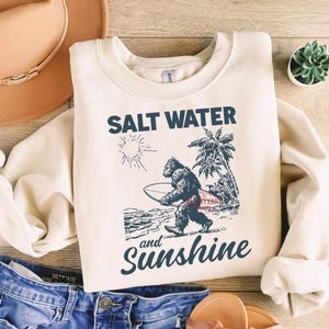 May include: Cream-colored sweatshirt with the words "SALT WATER and Sunshine" in navy blue script. A graphic of a Bigfoot-like creature carrying a surfboard is featured. The design includes a sun, palm trees, and ocean waves.
