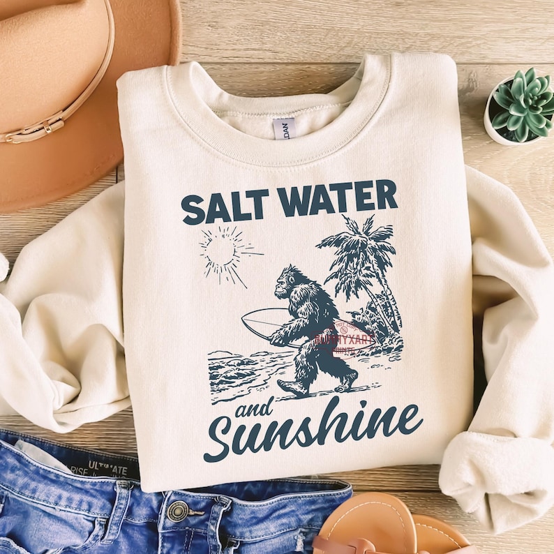 May include: Cream-colored sweatshirt with the words "SALT WATER and Sunshine" in navy blue script. A graphic of a Bigfoot-like creature carrying a surfboard is featured. The design includes a sun, palm trees, and ocean waves.