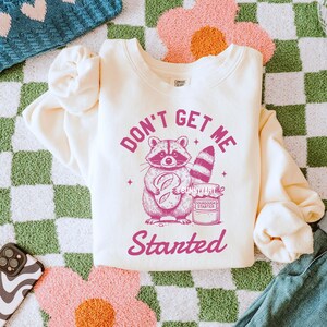 May include: A cream-colored sweatshirt with a pink graphic of a raccoon holding a loaf of bread and a jar of sourdough starter. The text "Don't Get Me Started" is printed in pink below the graphic.