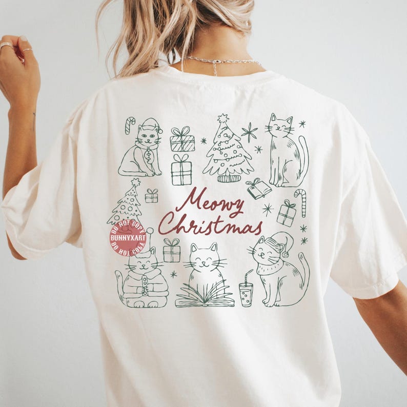 May include: Cream-colored t-shirt with a green line drawing of cats, Christmas trees, and gifts. The text "Meowy Christmas" is in burgundy script. The shirt has a relaxed fit.
