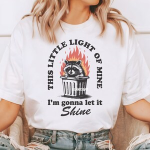 May include: White t-shirt featuring a raccoon in a trash can surrounded by flames. The text reads "THIS LITTLE LIGHT OF MINE I'm gonna let it Shine" in a circular and linear layout. The design includes the text "BunnyXart".