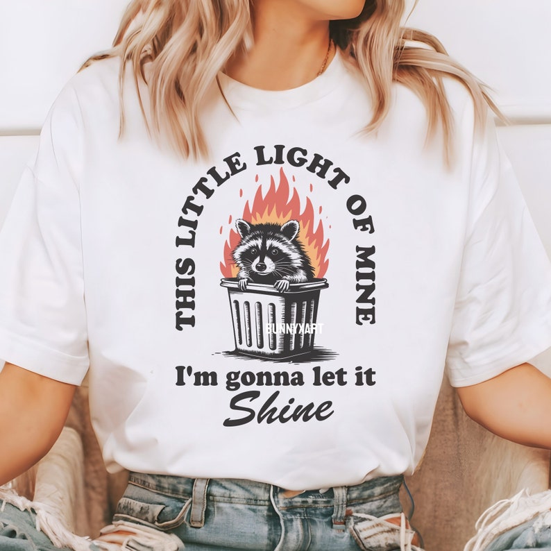 May include: White t-shirt featuring a raccoon in a trash can surrounded by flames. The text reads "THIS LITTLE LIGHT OF MINE I'm gonna let it Shine" in a circular and linear layout. The design includes the text "BunnyXart".