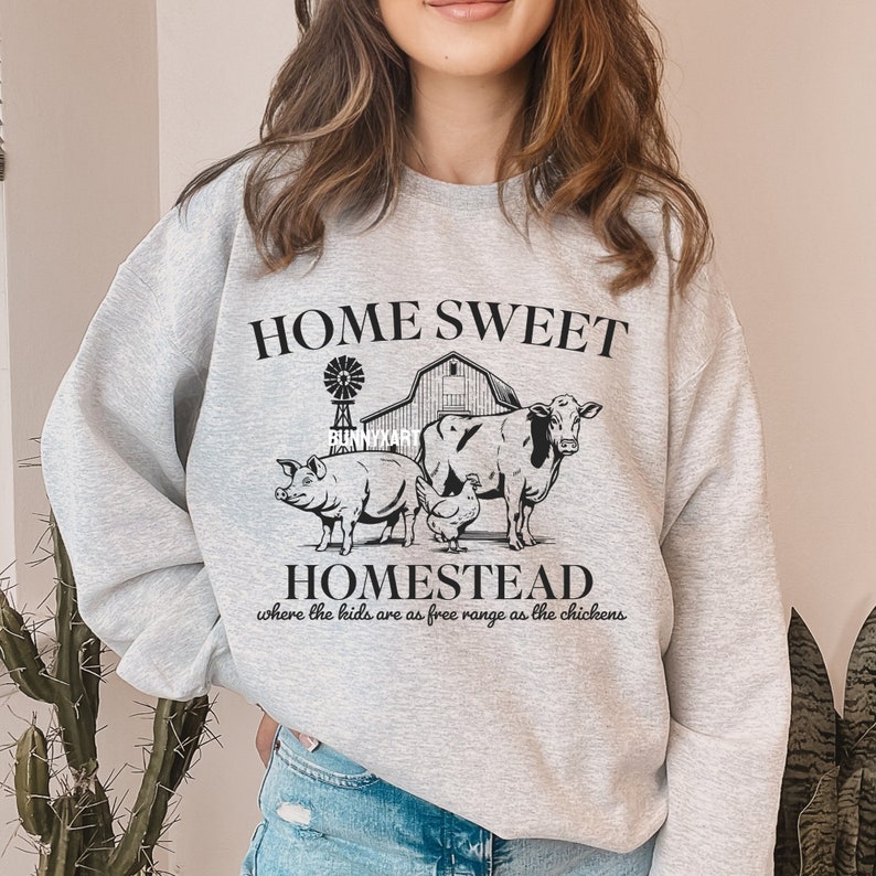 Pode incluir: Camisola cinzenta com o texto "Home Sweet Homestead" e um gr&aacute;fico de um celeiro, um moinho de vento, uma vaca, um porco e uma galinha. O texto abaixo do gr&aacute;fico diz "where the kids are as free range as the chickens".