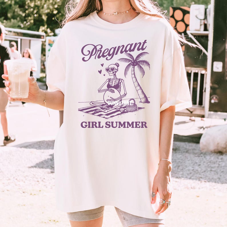 May include: A white t-shirt featuring a purple graphic of a skeleton wearing sunglasses and a bikini, sitting on a beach towel with a palm tree and the text "Pregnant Girl Summer".
