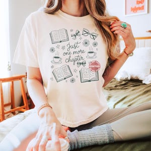 Op de afbeelding: Cr&egrave;mekleurig T-shirt met een grillig ontwerp met open boeken, brillen, strikken, bloemen en koffiekopjes. De tekst "Just one more chapter" is geschreven in een speels lettertype. Het shirt is een comfortabele, casual stijl.