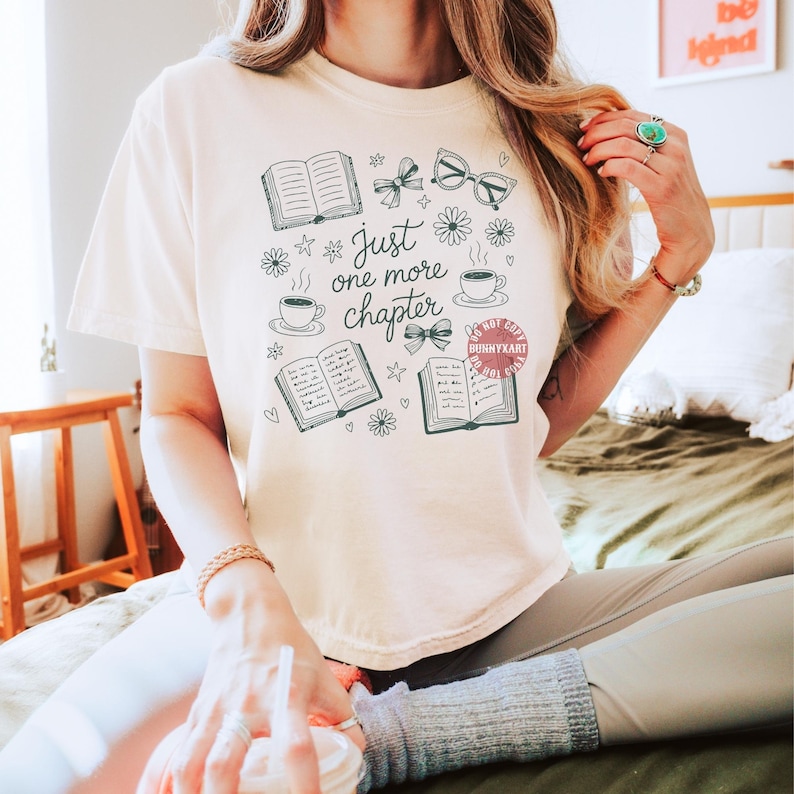 Op de afbeelding: Cr&egrave;mekleurig T-shirt met een grillig ontwerp met open boeken, brillen, strikken, bloemen en koffiekopjes. De tekst "Just one more chapter" is geschreven in een speels lettertype. Het shirt is een comfortabele, casual stijl.