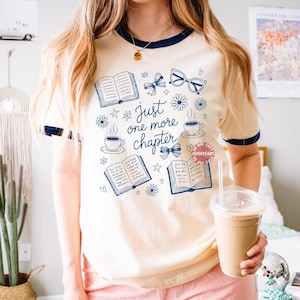 Op de afbeelding: Cr&egrave;mekleurig T-shirt met marineblauwe bies, met een ontwerp van boeken, bloemen, strikken, brillen en koffiekopjes. De tekst "Just one more chapter" staat in het midden van het ontwerp.