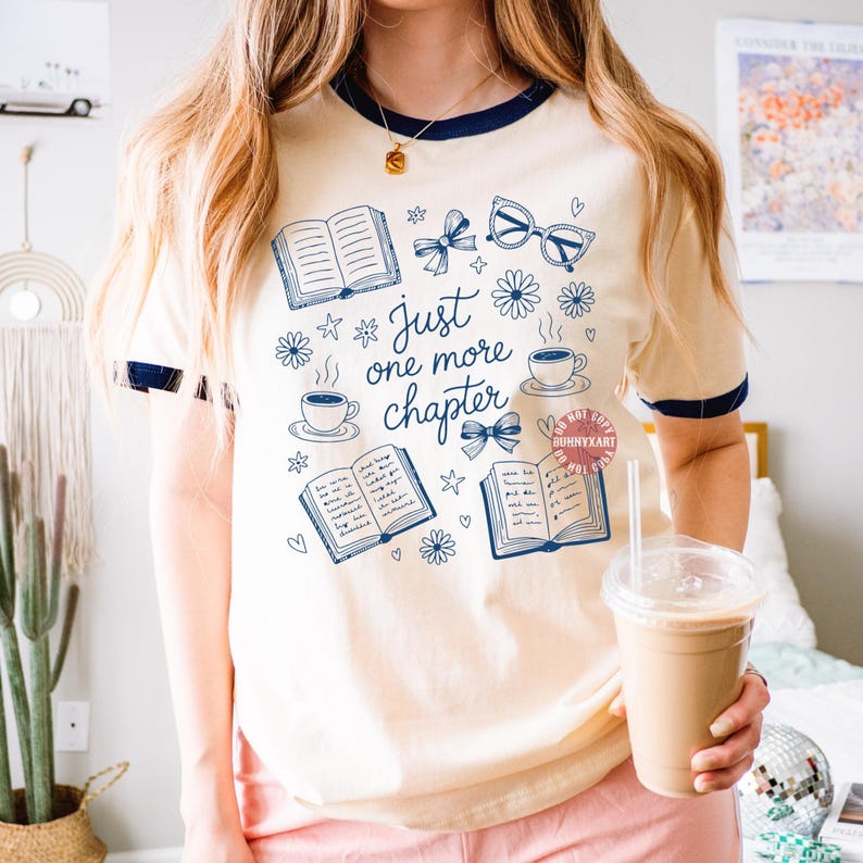 Op de afbeelding: Cr&egrave;mekleurig T-shirt met marineblauwe bies, met een ontwerp van boeken, bloemen, strikken, brillen en koffiekopjes. De tekst "Just one more chapter" staat in het midden van het ontwerp.