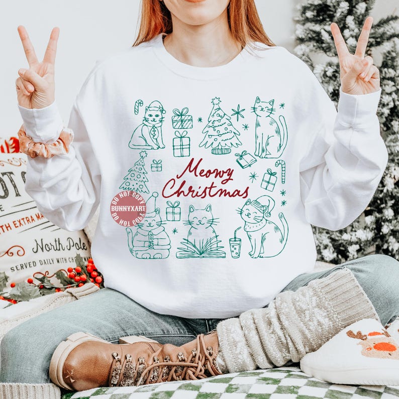 May include: White sweatshirt with a green and red "Meowy Christmas" design. The graphic features cartoon cats, Christmas trees, and gift boxes. The sweatshirt is a crew neck style.