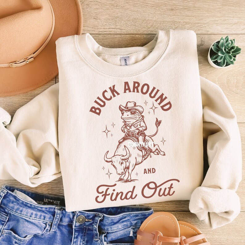 May include: A cream-colored sweatshirt with a brown frog riding a bull and the text "Buck Around and Find Out".