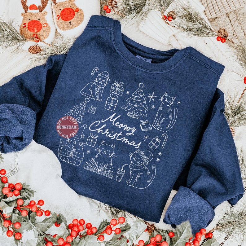 May include: Navy blue sweatshirt with a white graphic of cats, Christmas trees, and gifts. The text "Meowy Christmas" is also included in the design. The sweatshirt is surrounded by Christmas decorations.