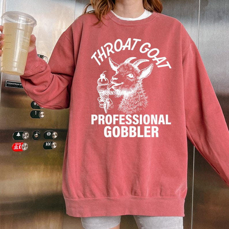 May include: A red sweatshirt with a white graphic of a goat eating an ice cream cone. The text on the sweatshirt reads "Throat Goat Professional Gobbler".