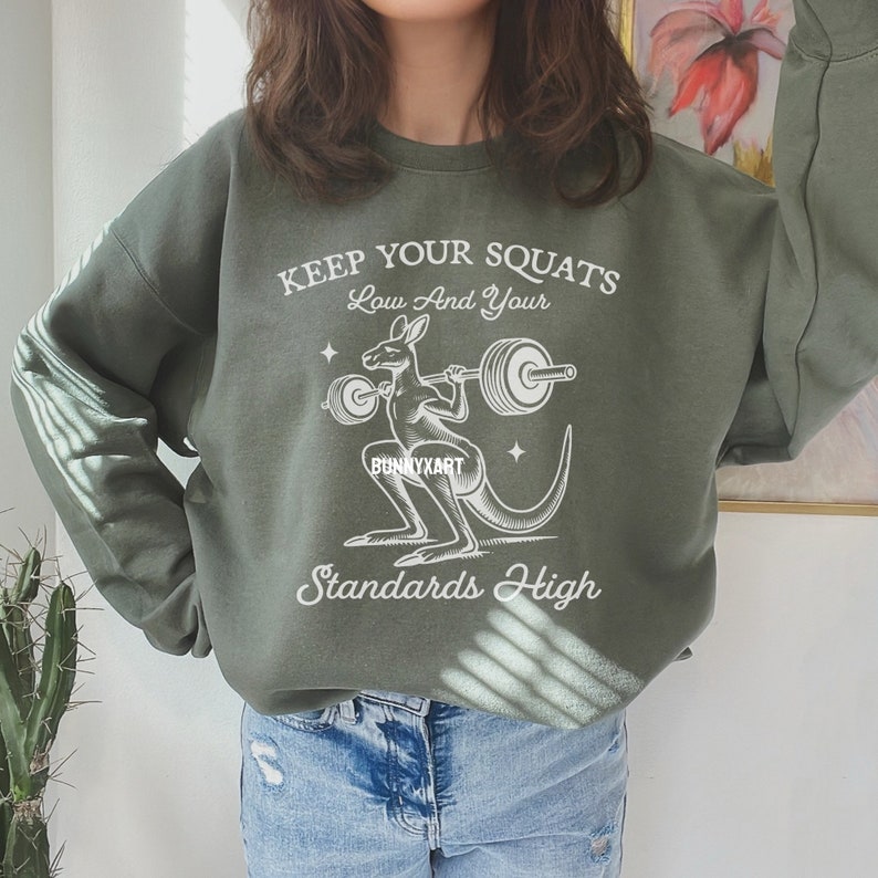 Pode incluir: Camisola verde com um gr&aacute;fico branco de um canguru a levantar uma barra. O texto na camisola diz "Keep your squats low and your standards high".