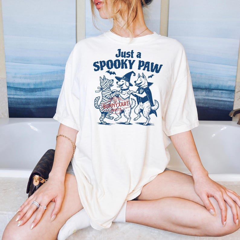 May include: Cream-colored t-shirt with a Halloween-themed graphic. The graphic features three dogs dressed as a mummy, a witch, and a vampire, with the text "Just a SPOOKY PAW". The shirt is a casual, oversized fit.