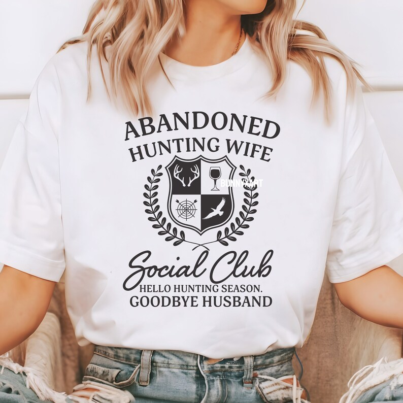 Op de afbeelding: Wit T-shirt met zwarte tekst die "Abandoned Hunting Wife Social Club Hello Hunting Season Goodbye Husband" luidt. De tekst wordt omringd door een zwart-wit wapenschild met een jachtthema.