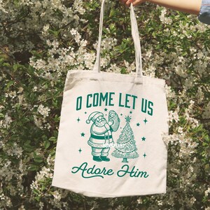 Pode incluir: Saco tote creme com uma ilustra&ccedil;&atilde;o verde do Pai Natal segurando um beb&eacute; ao lado de uma &aacute;rvore de Natal. O texto "O COME LET US Adore Him" est&aacute; impresso acima e abaixo da ilustra&ccedil;&atilde;o. O saco tem al&ccedil;as longas.