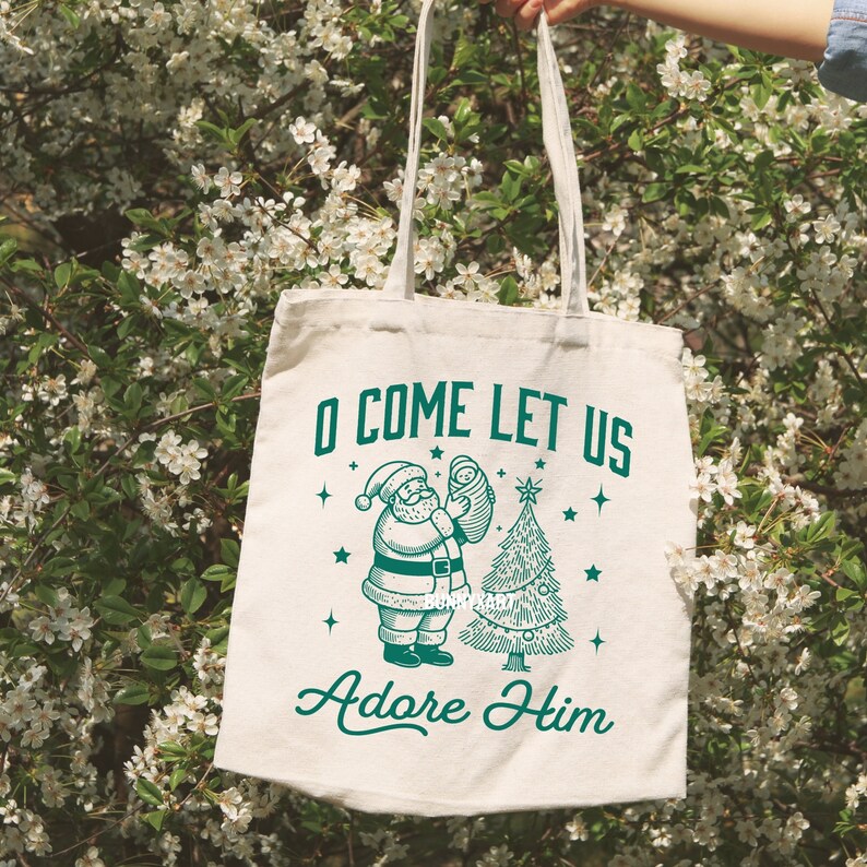 Pode incluir: Saco tote creme com uma ilustra&ccedil;&atilde;o verde do Pai Natal segurando um beb&eacute; ao lado de uma &aacute;rvore de Natal. O texto "O COME LET US Adore Him" est&aacute; impresso acima e abaixo da ilustra&ccedil;&atilde;o. O saco tem al&ccedil;as longas.