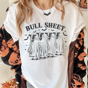May include: White t-shirt with the text "BULL SHEET" above a graphic of four cows dressed as ghosts, each holding a jack-o'-lantern. Bats and stars surround the cows. A black bat necklace is visible.