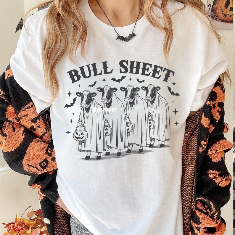 May include: White t-shirt with the text "BULL SHEET" above a graphic of four cows dressed as ghosts, each holding a jack-o'-lantern. Bats and stars surround the cows. A black bat necklace is visible.