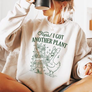 May include: Cream-colored sweatshirt with the text "Oops! I Got Another Plant" in green. A cartoon duck with a bow holds a potted plant, and a shelf with plants is also depicted. The sweatshirt is a casual, comfortable style.