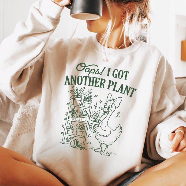 May include: Cream-colored sweatshirt with the text "Oops! I Got Another Plant" in green. A cartoon duck with a bow holds a potted plant, and a shelf with plants is also depicted. The sweatshirt is a casual, comfortable style.