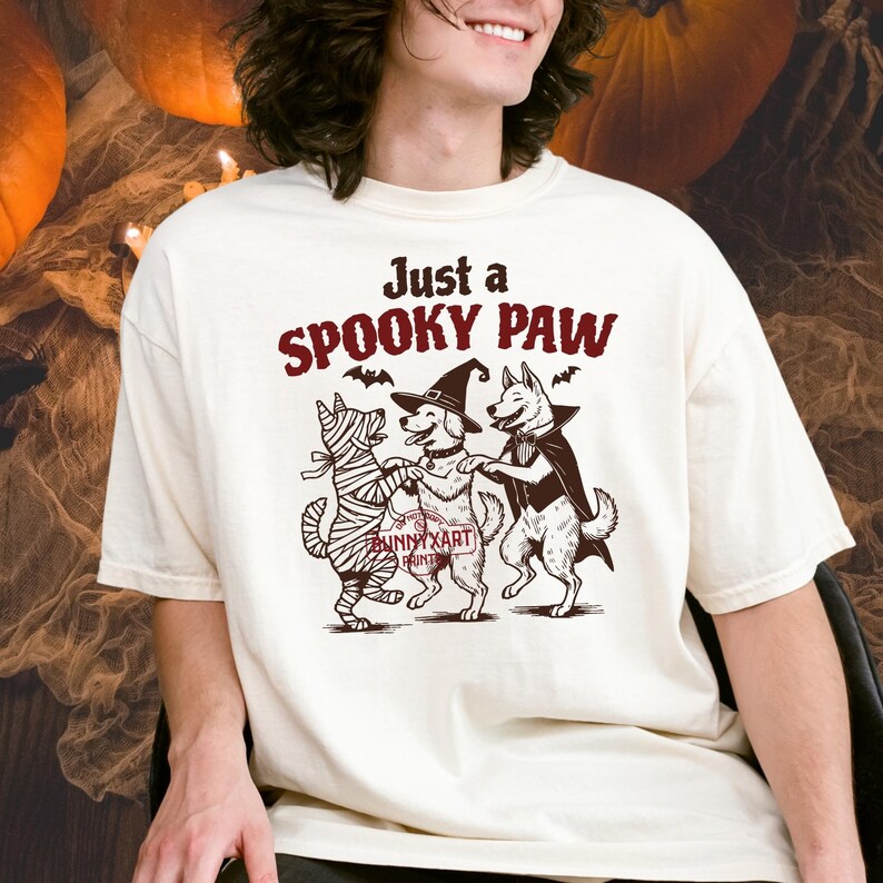 May include: Cream-colored t-shirt with a Halloween-themed graphic. The design features three dogs dressed in costumes: a mummy, a witch, and a vampire. The text "Just a Spooky Paw" is above the dogs.