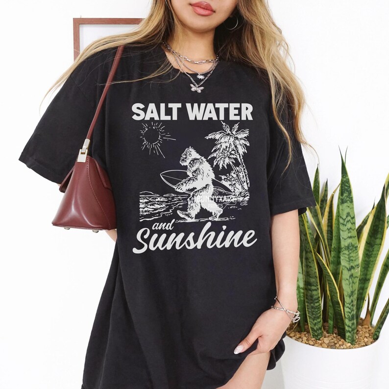 May include: Black t-shirt with the words "SALT WATER and Sunshine" in white. The graphic features a Bigfoot-like creature carrying a surfboard, palm trees, and a sun. The shirt is a casual, oversized style.