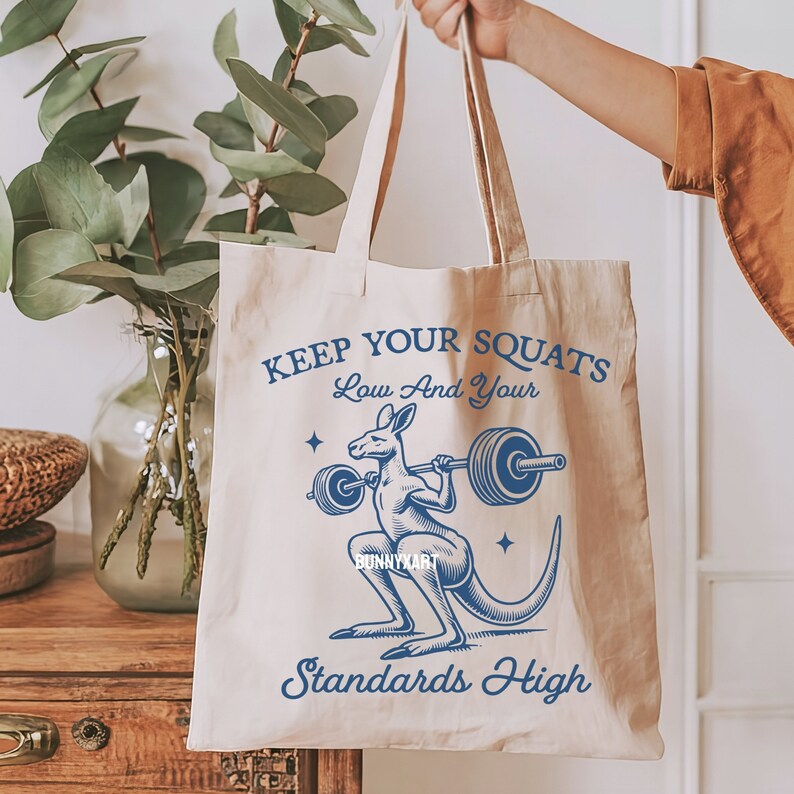 Pode incluir: Uma bolsa de tecido bege com uma ilustra&ccedil;&atilde;o azul de um canguru levantando uma barra de pesos. O texto na bolsa diz "Keep Your Squats Low And Your Standards High".