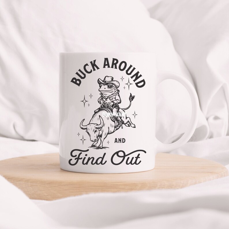 May include: White ceramic mug with a black and white illustration of a frog riding a bull. The text "Buck Around and Find Out" is printed on the mug.