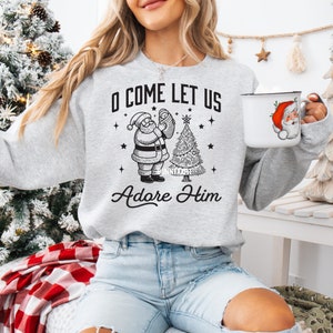 Pode incluir: Um moletom cinza claro com um gr&aacute;fico em preto e branco do Papai Noel segurando um beb&ecirc; ao lado de uma &aacute;rvore de Natal. O texto "O COME LET US Adore Him" est&aacute; impresso no moletom. Uma caneca branca com um desenho do Papai Noel tamb&eacute;m &eacute; vis&iacute;vel.