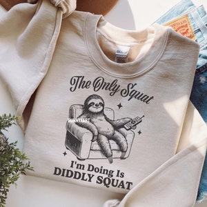 May include: A beige sweatshirt featuring a graphic of a sloth relaxing in an armchair with a remote control. The text on the sweatshirt reads "The Only Squat I'm Doing Is Diddly Squat."