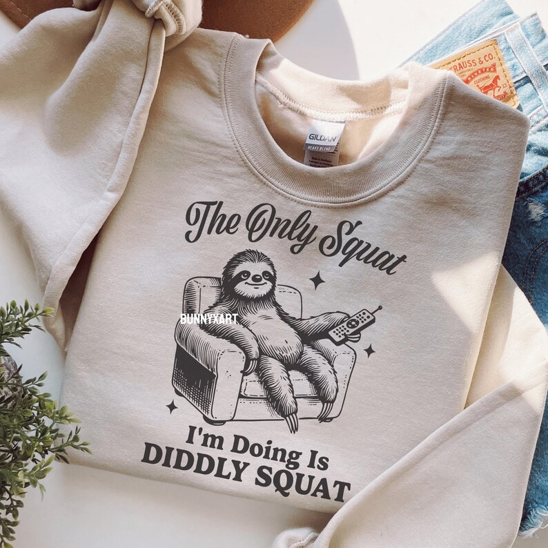 May include: A beige sweatshirt featuring a graphic of a sloth relaxing in an armchair with a remote control. The text on the sweatshirt reads "The Only Squat I'm Doing Is Diddly Squat."