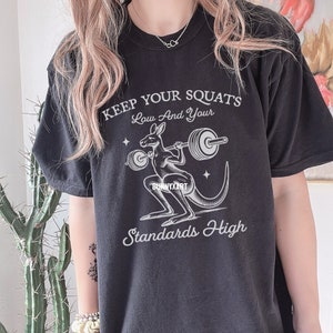Pode incluir: Camiseta preta com um gr&aacute;fico branco de um canguru levantando pesos. O texto "Keep Your Squats Low And Your Standards High" est&aacute; impresso na camiseta.