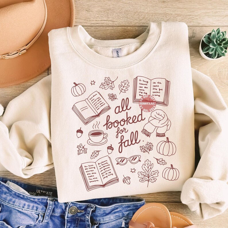 Może przedstawiać: Kremowa bluza z nadrukiem w jesiennym klimacie. Projekt zawiera dynie, liście, książki, filiżankę kawy i napis "all booked for fall". Bluza leży na niebieskich dżinsach i brązowym kapeluszu.