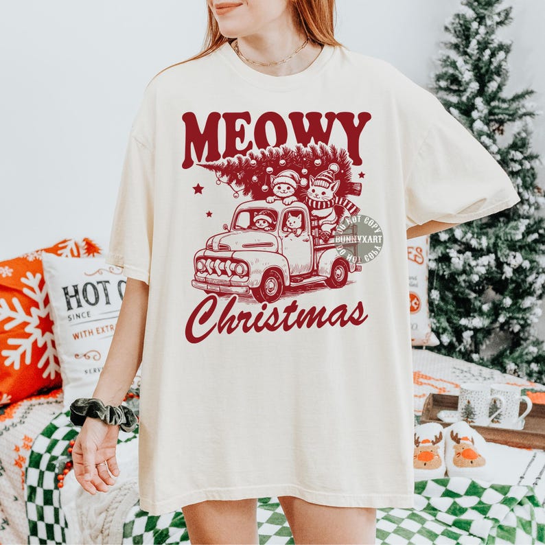 May include: Cream-colored t-shirt with a red graphic featuring a vintage truck carrying a Christmas tree and cats. The text "MEOWY Christmas" is arched above the truck. A festive holiday design.
