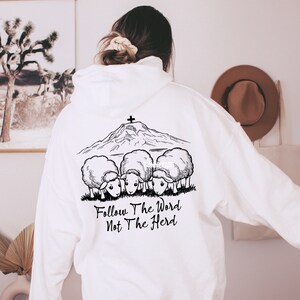 May include: White hoodie with a black and white graphic of a mountain with a cross on top and four sheep looking at the mountain. The text "Follow The Word Not The Herd" is printed below the graphic.