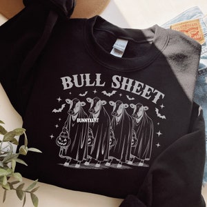 May include: Black sweatshirt with the text "BULL SHEET" above a graphic of four cows dressed as ghosts, each holding a jack-o'-lantern. Bats and stars surround the cows. The brand name "BUNNYXART" is at the bottom.