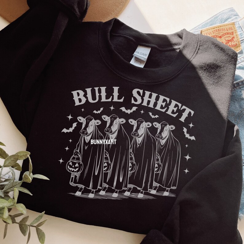 May include: Black sweatshirt with the text "BULL SHEET" above a graphic of four cows dressed as ghosts, each holding a jack-o'-lantern. Bats and stars surround the cows. The brand name "BUNNYXART" is at the bottom.