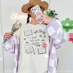 Op de afbeelding: Cr&egrave;mekleurig T-shirt met een zwarte afbeelding van boeken, strikken, brillen en koffiekopjes. De tekst "Just one more chapter" staat erop. De persoon draagt een lichtpaars en wit geruit vest en een strohoed.