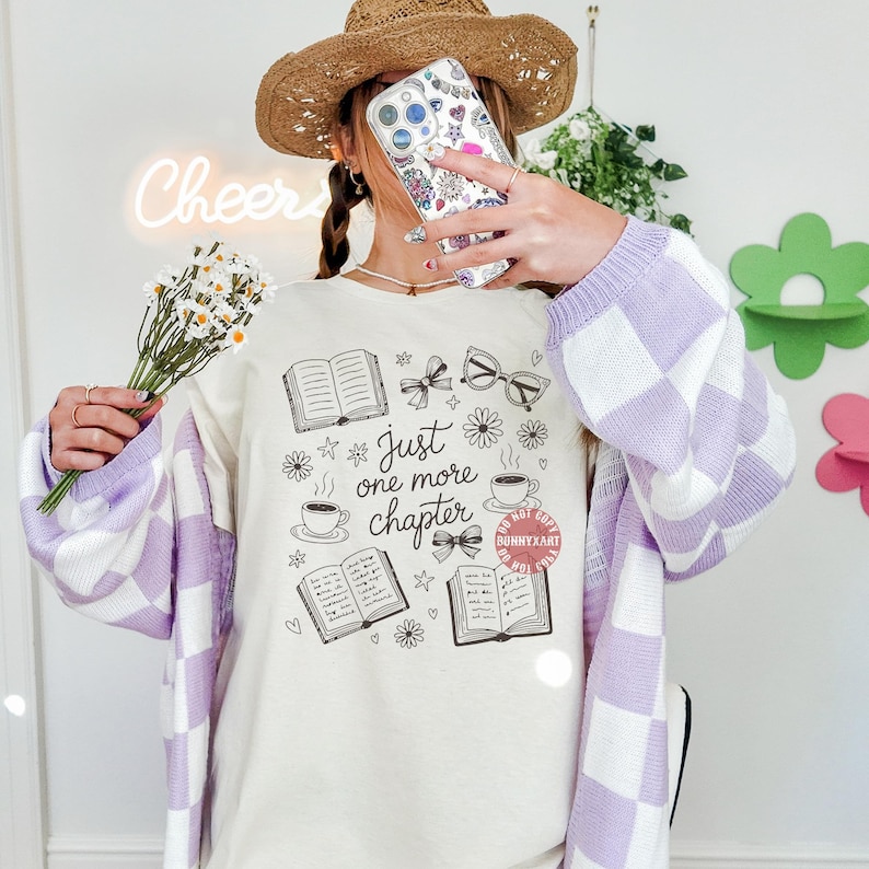 Op de afbeelding: Cr&egrave;mekleurig T-shirt met een zwarte afbeelding van boeken, strikken, brillen en koffiekopjes. De tekst "Just one more chapter" staat erop. De persoon draagt een lichtpaars en wit geruit vest en een strohoed.