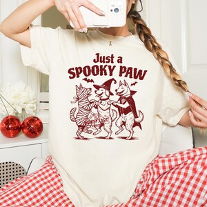 May include: Cream-colored t-shirt with a Halloween-themed graphic. The graphic features three dogs dressed as a mummy, a witch, and a vampire, with the text "Just a Spooky Paw". Bats are also included in the design.