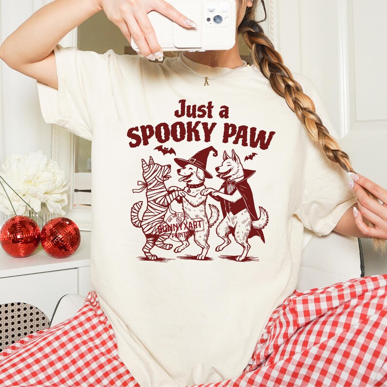May include: Cream-colored t-shirt with a Halloween-themed graphic. The graphic features three dogs dressed as a mummy, a witch, and a vampire, with the text "Just a Spooky Paw". Bats are also included in the design.