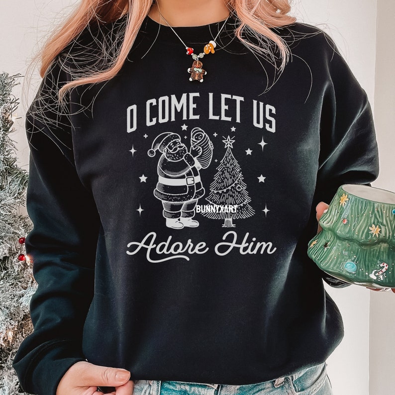 Pode incluir: Camisola preta com um gr&aacute;fico branco apresentando o Pai Natal segurando um beb&ecirc;, uma &aacute;rvore de Natal e o texto "O Come Let Us Adore Him". O design inclui estrelas e a assinatura do artista.