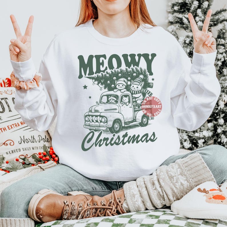May include: White sweatshirt with a green graphic of a vintage truck carrying a Christmas tree and cats. The text reads "MEOWY Christmas". The sweatshirt is worn by a person making a peace sign.