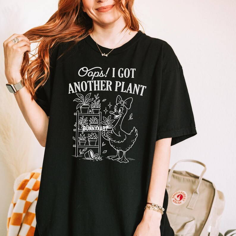 May include: Black t-shirt with the text "Oops! I Got Another Plant" in white. The shirt features a white graphic of a duck with a bow, a plant shelf, and the text "Bunnyart".