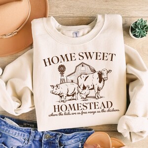 Pode incluir: Uma camisa de moletom cor creme com o texto "HOME SWEET HOMESTEAD where the kids are as free range as the chickens" impresso em marrom. O gr&aacute;fico apresenta um celeiro, um moinho de vento, uma vaca, um porco e uma galinha.