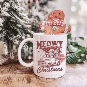 May include: A white ceramic mug with the words "MEOWY Christmas" in maroon lettering. The mug features a graphic of a vintage truck carrying a Christmas tree and cats. A gingerbread man cookie sits atop the mug.