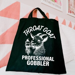 May include: Black tote bag with a white graphic of a goat eating ice cream. The text on the bag reads "Throat Goat Professional Gobbler".